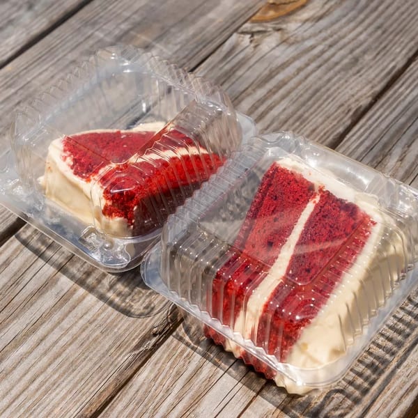 Two slices of red velvet cake with cream cheese frosting in clear plastic containers on a wooden table.