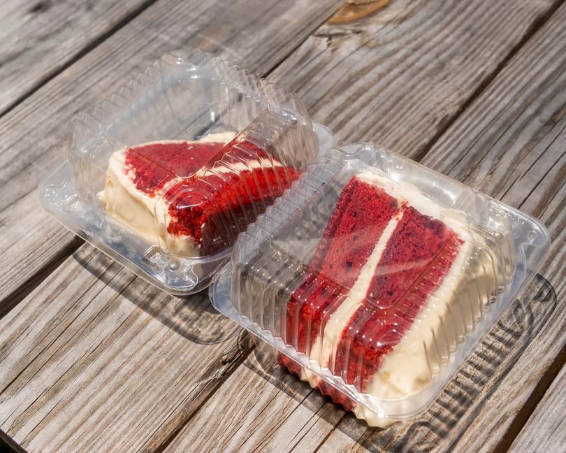 Two slices of red velvet cake with cream cheese frosting in clear plastic containers on a wooden table.