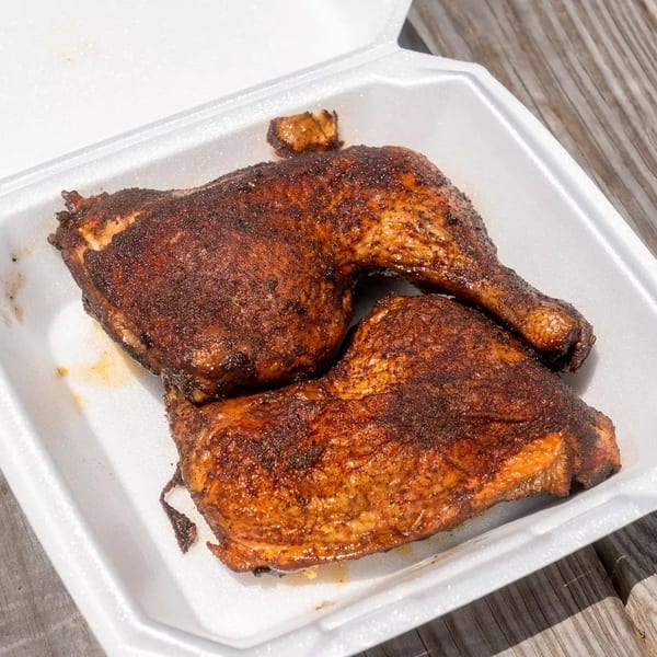 Two seasoned, dark brown grilled chicken leg quarters in a white takeout container on a wooden table.