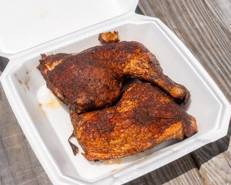 Two seasoned, dark brown grilled chicken leg quarters in a white takeout container on a wooden table.