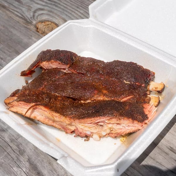 Smoked ribs with a dark rub in a white takeout container on a wooden table.