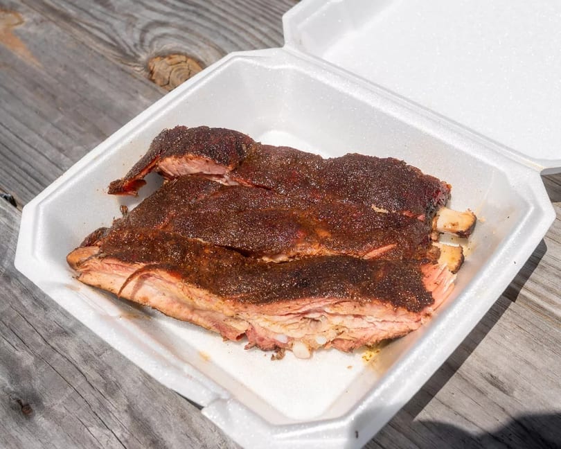 Smoked ribs with a dark rub in a white takeout container on a wooden table.