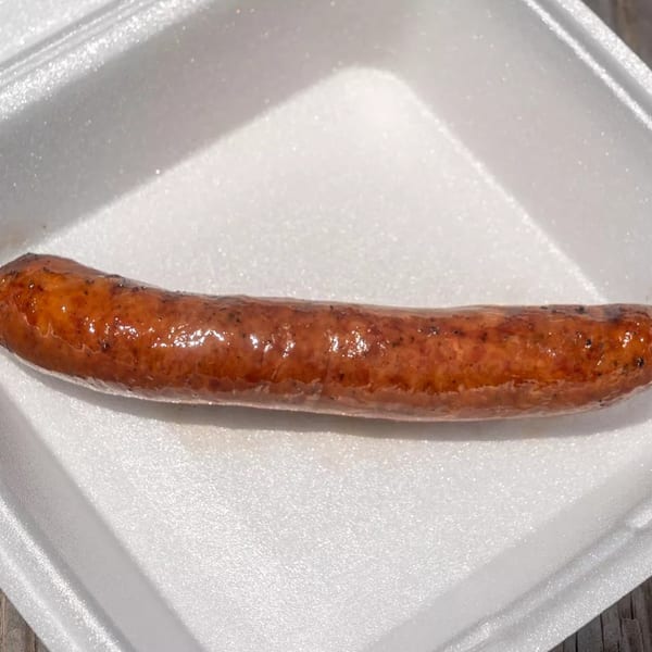 A single grilled sausage with a shiny, browned casing rests in a white styrofoam container.