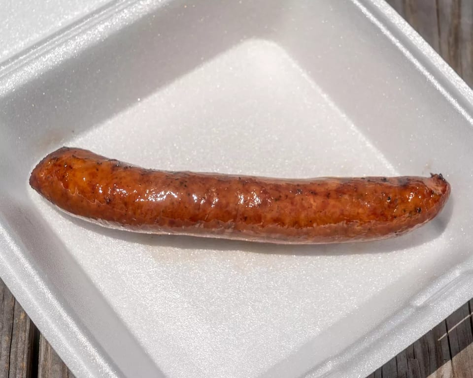 A single grilled sausage with a shiny, browned casing rests in a white styrofoam container.
