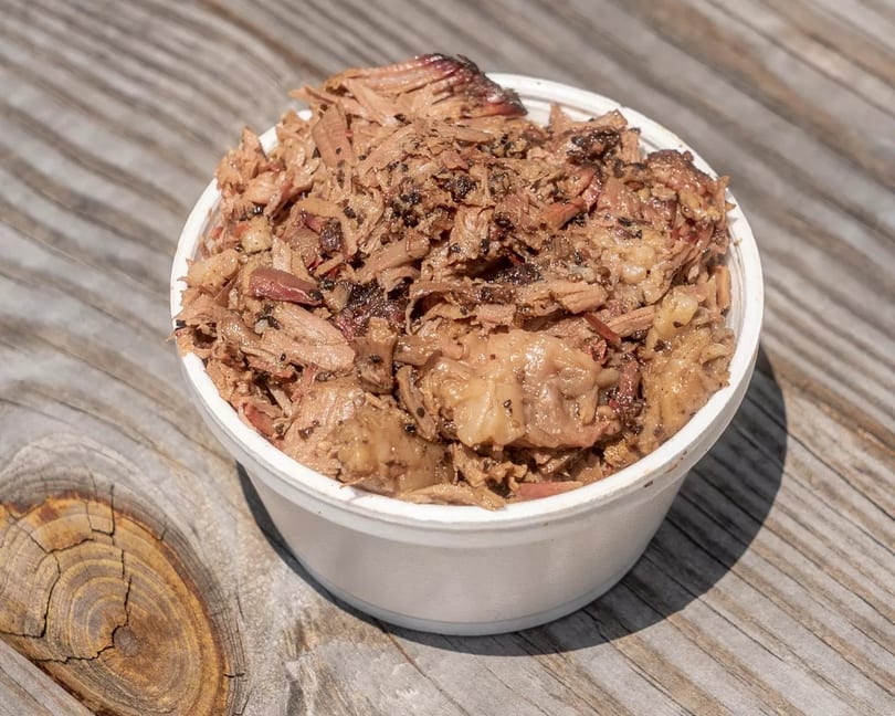 A white bowl filled with shredded, seasoned barbecue meat sits on a rustic wooden surface.