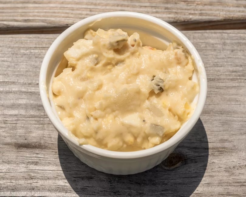Creamy potato salad with visible chunks of potato and relish in a white bowl on a wooden surface.
