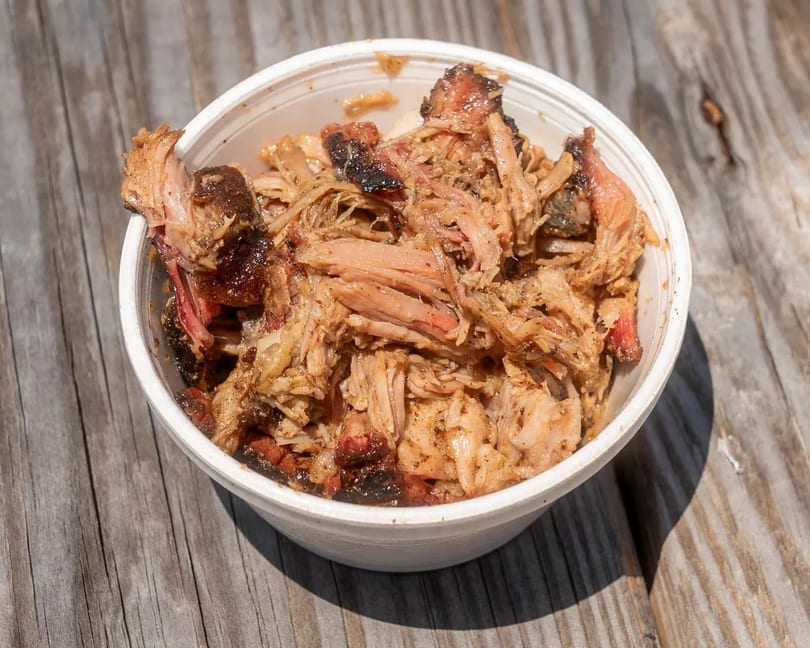 A white bowl filled with juicy pulled pork, showing crispy edges and tender meat, on a wooden table.